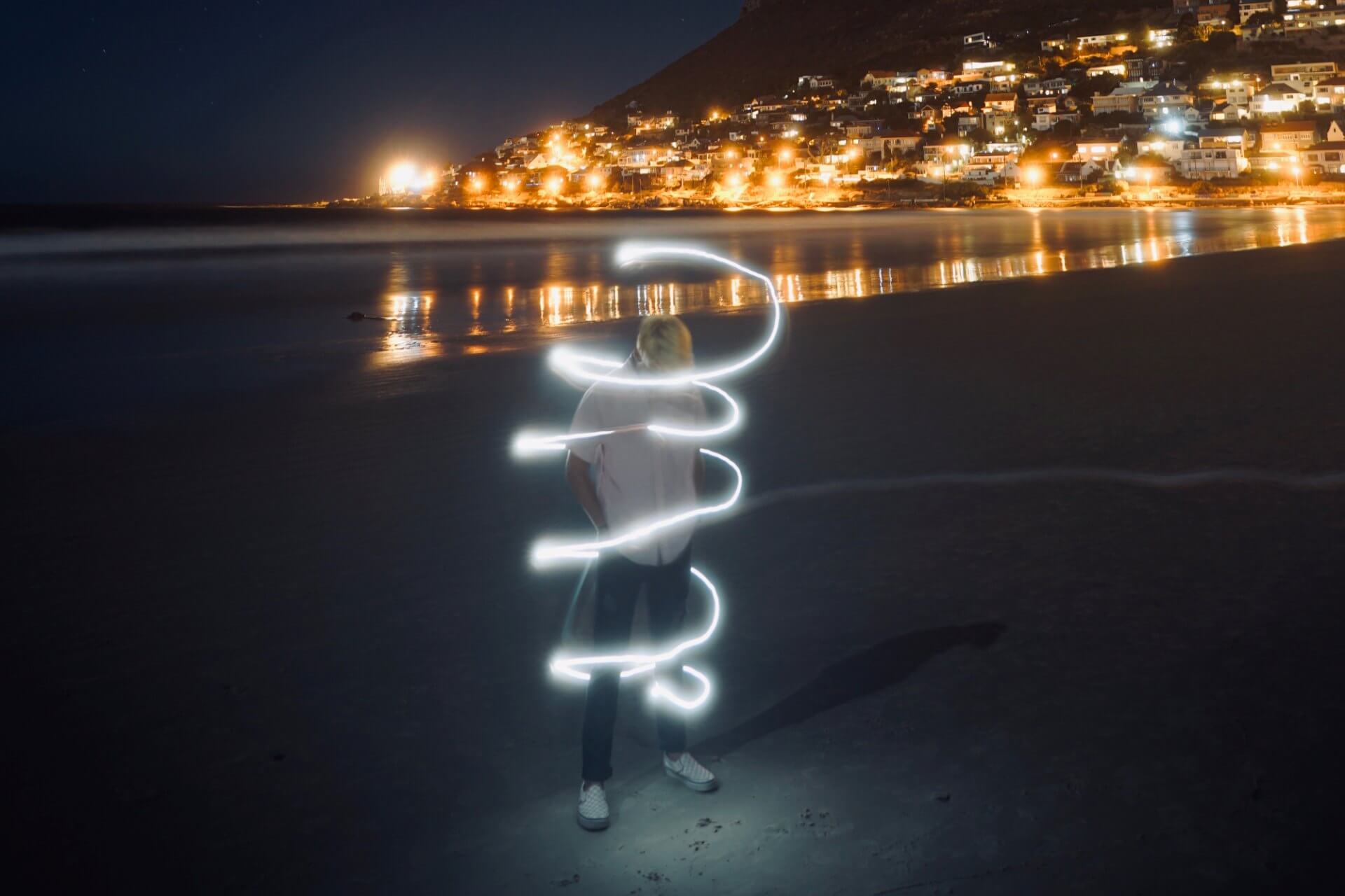 A man stands on a beach in Cape Town at night with a swirling light around him while street lights illuminate the water from across the bay. (Photo by Clayton Cardinalli on Unsplash)