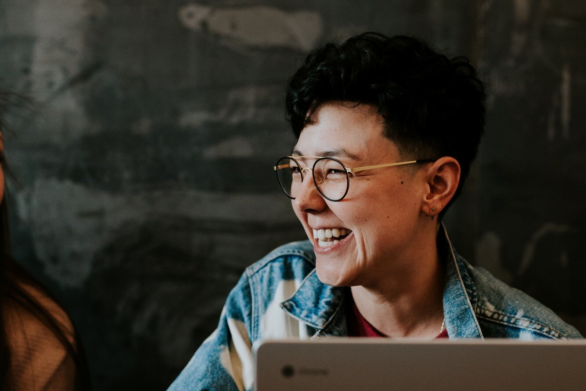 A woman in a denim jacket and glasses sitting at her laptop smiles broadly (Photo by Brooke Cagle on Unsplash)
