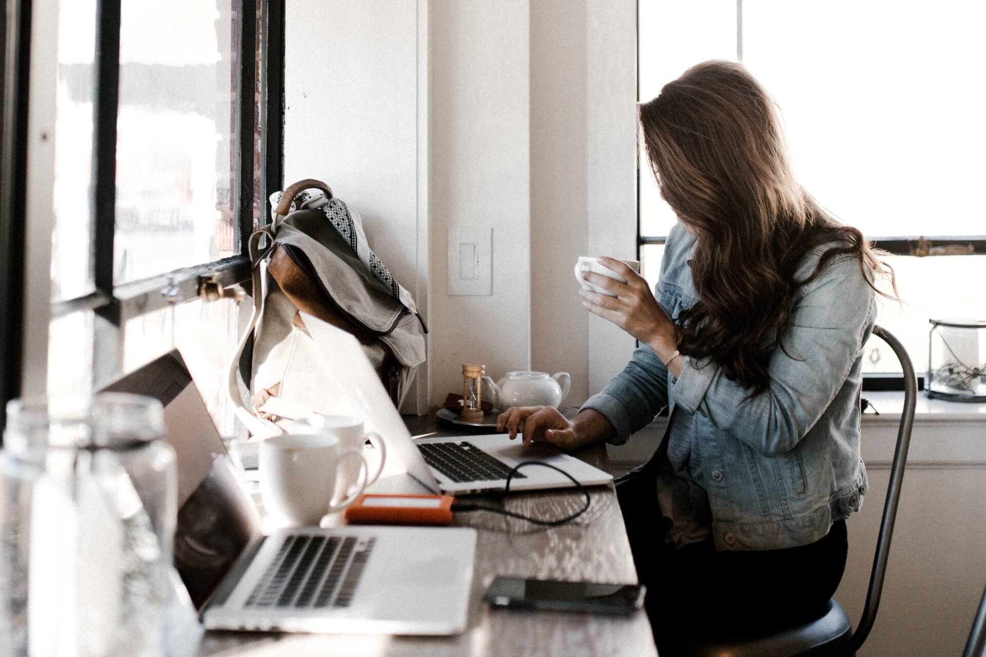 A woman with long brown hair wearing a denim jacket works at her laptop while drinking a cup of tea (Photo by Andrew Neel on Unsplash)