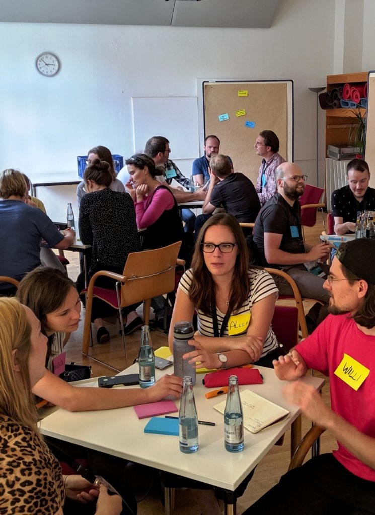 Jock Busuttil runs a product management training workshop for a room of people with name tags seated at square tables at Working Products 2019 (Photo by Jock Busuttil / Product People Limited)