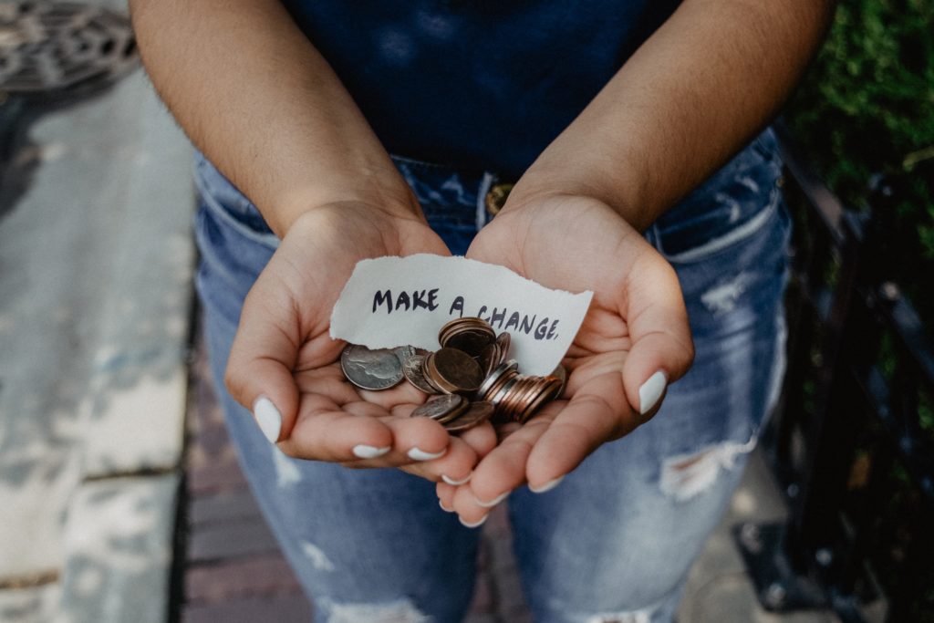 A woman holds spare change in her cupped hands with a note saying "Make a change" (Photo by Kat Yukawa on Unsplash)