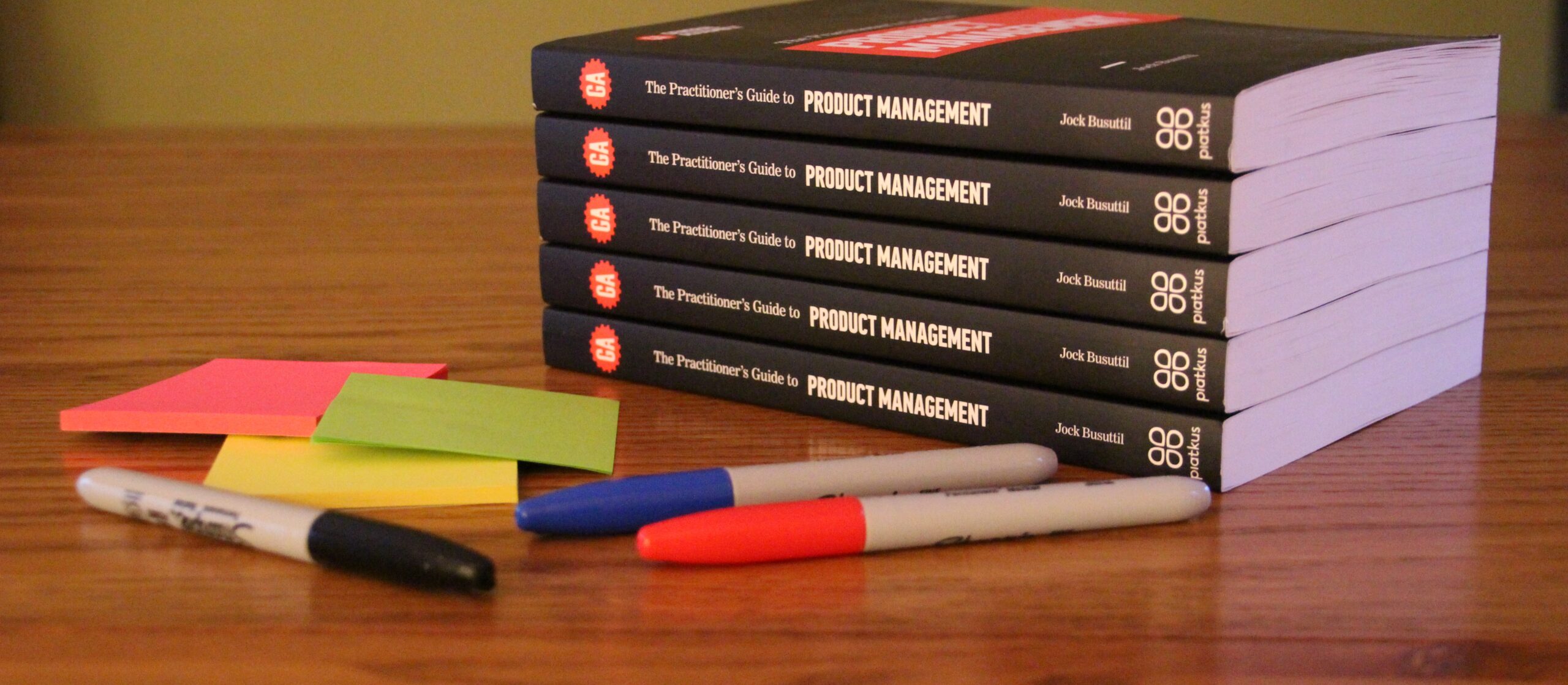 A pile of books "The Practitioner's Guide to Product Management" sits on a table next to some colourful sticky notes and Sharpie pens (Photo by Jock Busuttil / Product People Limited)
